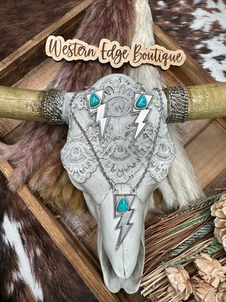 A decorative cow skull adorned with the Hazer Turquoise Lighting Bolt Set is displayed against a rustic wooden backdrop. The background features cowhide patterns and a wooden sign that reads "Western Edge Boutique," completing the perfect Western outfit.