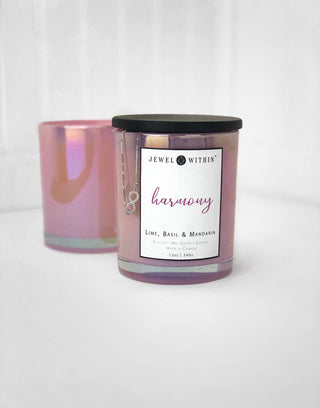 A "Harmony Jewelry Candle" from "Jewel Within," featuring a lime, basil, and mandarin scent, is in focus. The iridescent pink jar with a black lid holds an eco-friendly candle made from natural coconut wax, while a silver necklace draped over it adds elegance. Another similar candle jar is blurred in the background.