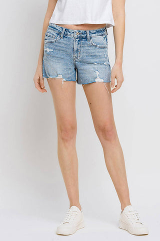 A person is wearing Kylie Mid Rise Raw Distressed Hem Shorts from VERVET by FLYING MONKEY and a white cropped top, paired with white sneakers. The shorts, made from comfortable stretch denim, have a slightly frayed hem and visible wear marks for a casual look. The individual is standing against a plain light-colored background.