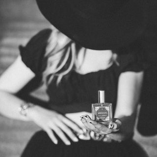 Black and white photo of a person with long hair, wearing a wide-brimmed hat and holding a small bottle labeled "Midnight Perfume" by R. Rebellion. Their face is partially obscured by the hat, and they are dressed in a dark, flowing outfit. This daring fragrance promises an enchanting fruity-floral scent.