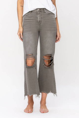 A person stands barefoot against a plain backdrop, wearing a white top and VERVET by Flying Monkey's Kutter Metal Gray Distressed Vintage Crop Flare Jeans. The non-stretch denim jeans are distressed with large rips on both knees and have frayed hems that end slightly above the ankles.