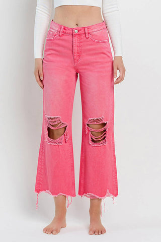 A person is standing barefoot in bright pink, Rosie 90's Super High Rise Crop Flare Hot Pink Jeans by VERVET by FLYING MONKEY, featuring wide legs with ripped knees and frayed hems that are reminiscent of 90's vintage jeans. They are wearing a long-sleeve white top, cropped at the waist. The background is plain white.