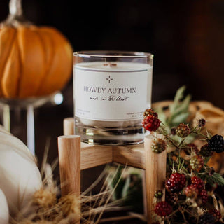 An exclusive "Howdy Autumn Candle" by R. Rebellion, 8 oz., rests on a wooden stand surrounded by fall decor, including a small pumpkin and a cluster of berries. The Wild West-inspired backdrop is softly blurred, accentuating the candle and its cozy seasonal setting.