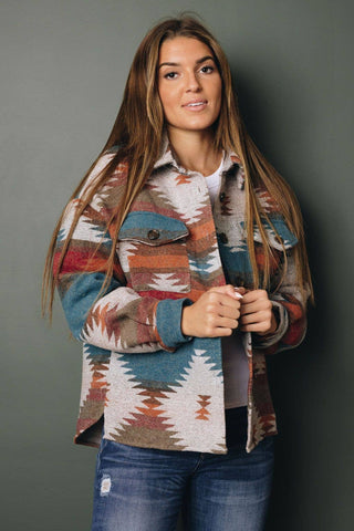 A woman with long brown hair stands against a gray-green background, wearing the Zayn Aztec Jacket by Stay Warm in Style, made of 100% polyester with a southwestern pattern in earthy tones over a true-to-size white top and blue jeans. She gazes at the camera neutrally.