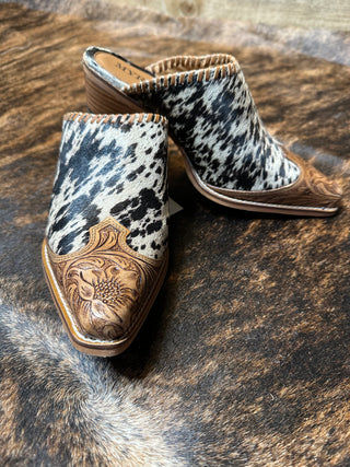 A pair of stylish Jennings Cowhide and Tooled Leather Mules by Western Edge Boutique with a pointed toe rests on a fur rug. These fashionable women's mules feature a striking mix of black-and-white cowhide pattern and intricate brown leather detailing on the toe and heel, making them the perfect complement to your favorite skinny jeans. Their medium-height stacked heel adds to their chic appeal.