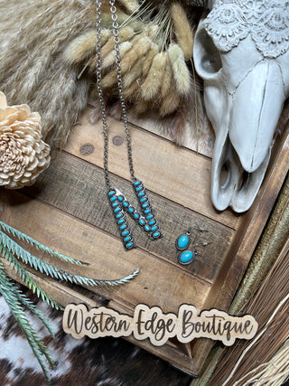 The Nattie “N” Letter Turquoise Necklace Earring Set, featuring a turquoise-studded "N" pendant and matching earrings crafted in brushed silver, is displayed on a rustic wooden surface. Surrounding items include a decorative animal skull, dried flowers, and a Western Edge Boutique sign.