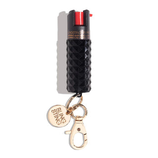 The Emergency Pepper Spray Key Ring by BLINGSTING is a chic black, textured canister with a red trigger and a gold keychain attachment. This stylish accessory includes UV marking dye and the attached keychain features a lobster clasp and a gold circular tag engraved with "BLINGSTING," making it part of a renowned women's safety brand.