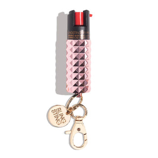 A pink, diamond-patterned canister of the Emergency Pepper Spray Key Ring features a black nozzle and red button on top. It comes with a gold keychain attachment featuring a lobster clasp and a circular tag engraved with "BLINGSTING," proudly representing the women's safety brand. The spray is equipped with UV marking dye for added security.