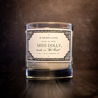 A lit candle in a clear glass jar sits on a dark surface. The label reads "R. Rebellion - Miss Dolly Candle 8 oz. - made with coconut soy and phthalate-free fragrance oils - Pendleton, OR" with decorative borders. The background is a textured dark brown, enhancing its luxurious blend.