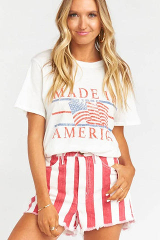 A person with long blonde hair is wearing a white t-shirt featuring the “Made in America” slogan and an American flag graphic. They complement this look with Freedom Ring Striped Distressed Shorts from western edge boutique, which are adorned with red and white vertical stripes and feature convenient front pockets. The background is plain white.