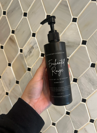 A hand holding a bottle of luxurious hand soap from Mavwicks Fragrances LLC, labeled "Swindler," against a backdrop of black, white, and gray hexagon tiles. Featuring a pump dispenser, the label highlights it as "Luxury & Modern" and includes natural oils for added skincare benefits.