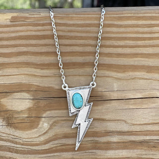 The Hallie Turquoise Stone Lightning Bolt Necklace by Wild Junkie features a silvertone pendant with a turquoise stone in the center of the bolt's upper section. The necklace is displayed on a wooden surface with visible grain patterns.