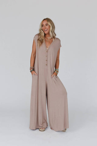 A woman with long blonde hair stands against a plain white background. She is wearing the Kendrick Mocha Jumpsuit from Three Bird Nest, a loose boho chic piece in light brown with a deep V-neck and buttons down the front. She accessorizes with bracelets on both wrists and smiles with her hands in the jumpsuit's pockets.