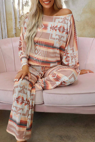 A person with long blonde hair is sitting on a light pink upholstered couch, wearing the Maxie Aztec Lounge Set by Lovesoft. The coordinated lounge set features an off-shoulder top and comfortable drawstring waist pants, both adorned with an Aztec print in shades of peach, beige, and white.