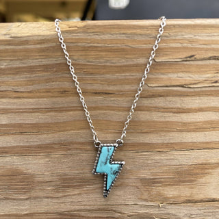 The Lindy Lightning Bolt Turquoise Necklace by Western Edge Boutique features a silver chain with a turquoise stone pendant, surrounded by a rugged, textured metal border. Perfect for accessorizing a western-inspired outfit, it is beautifully displayed on a wooden surface.