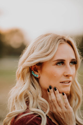 A woman with long wavy blonde hair is looking thoughtfully into the distance. She has her right hand near her chin, showcasing her black painted nails, and she is wearing the Harriet Turquoise Ear Climber Earring Set by Western Edge Boutique. The background is softly blurred.