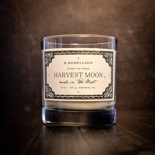 A candle jar labeled "R. Rebellion, Harvest Moon Candle," containing all-natural coconut soy wax and marked as 8 oz (227 g), made in Pendleton, OR. The jar is half-full and features an ornate, vintage-style decorative border against a dark, textured background.