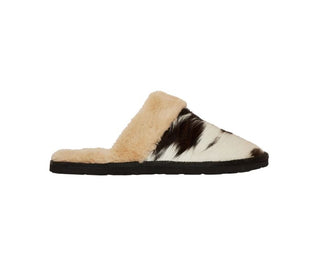 A side view of the Montana House Slipper from Western Edge Boutique, showcasing a snug slip-on mule with a fuzzy, cream-colored interior and a distinctive black and brown cowhide-patterned exterior. The slipper is equipped with a black rubber sole, ensuring both comfort and durability with its feathery texture.