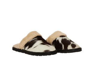 The Montana House Slipper from Western Edge Boutique features cowhide uppers with a black and white cow print design, tan-colored faux fur lining around the edges, and sturdy black rubber soles. Reminiscent of snug slip-on mules, these slippers are elegantly positioned side by side on a pristine white background.